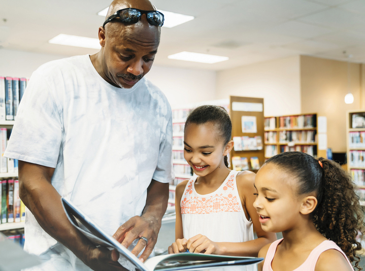 Black father and two daughters look at a book together in the library.