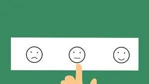 A piece of paper displays a sad face, a neutral face, and a smiley face as rating options. A human hand tentatively hovers over the neutral face.