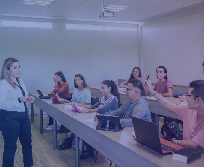 Female instructor leading a class of multiethnic young adults sitting classroom style with laptops and other mobile computing devices in front of them.