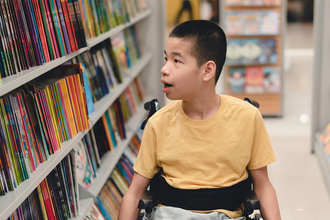 Asian child in a wheelchair looking a shelves of many books.