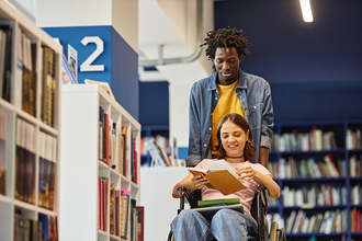 Young BIPOC male assisting a young female, who is in a wheelchair and holding books, in a library.